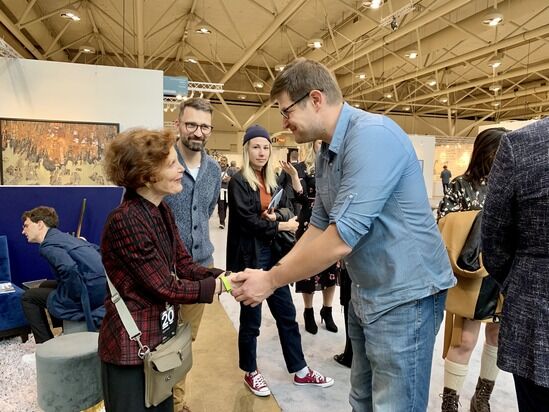 A man in a blue shirt shakes hands with an celebrated woman in a red and black plaid jacket at a busy indoor event. Several people are standing nearby, engaging in conversation. The setting appears to be a convention or exhibition hall with high ceilings and displays.