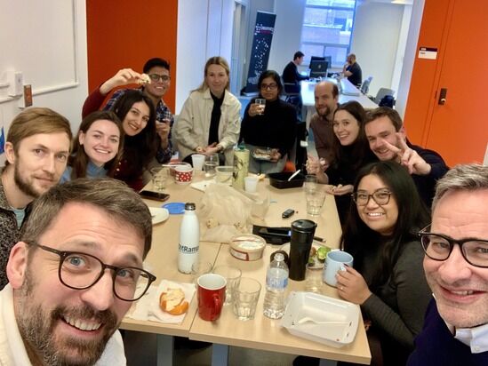 A group of eleven people sits around a cluttered table, smiling and posing for the camera in a brightly lit office setting. Some hold food and drinks, indicating a casual meal or get-together. A man in the foreground takes a selfie of the group.