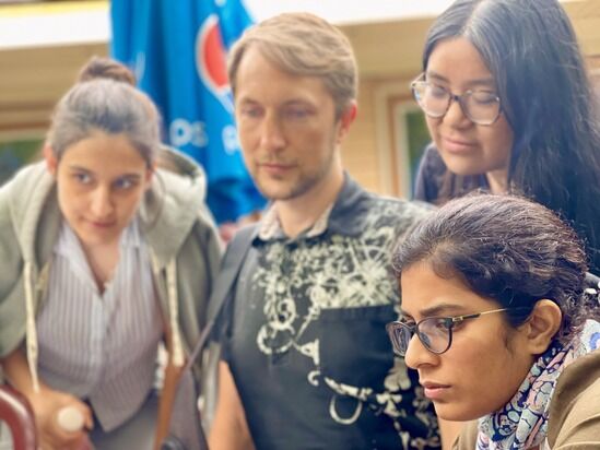 A group of four people, two women and one man with another woman visible partially, are closely gathered outdoors. They appear to be focused on something in front of them. A Pepsi umbrella is in the background.