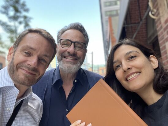 Three people, two men and one woman, smile at the camera while standing outside on a street with buildings and trees in the background. One man has glasses, and the woman holds an orange book.