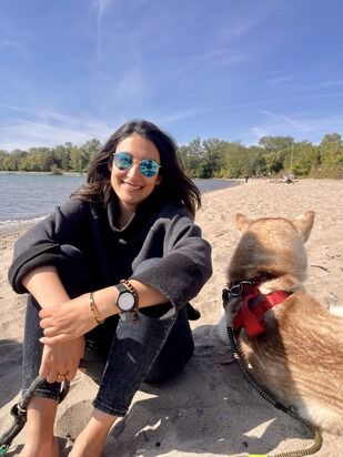A woman with long hair and sunglasses sits on a sandy beach, smiling at the camera. Beside her, a dog with a red harness looks away towards the water. The background features a calm lake and lush green trees under a clear blue sky.