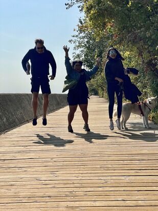 Three people are jumping mid-air on a wooden boardwalk surrounded by foliage. A person wearing black is on the left, the person in the middle is wearing dark clothes with a green jacket, and the person on the right is wearing dark clothes and a mask, holding a dog on a leash.