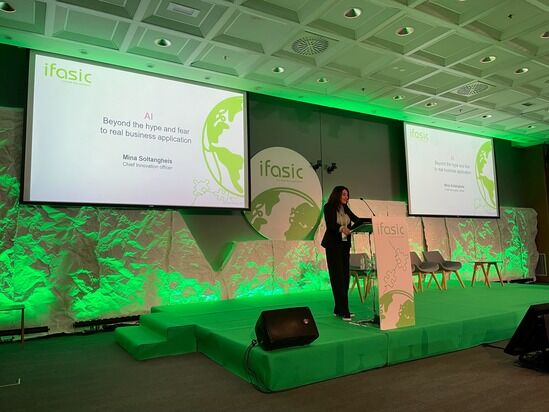 A speaker stands at a podium with a presentation slide behind them. The slide reads, 'AI Beyond the hype and fear to real business application.' The backdrop features green lighting and the logo ifasic.