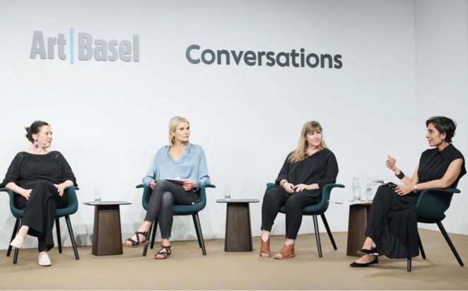 Four women sit and engage in a discussion on a stage during an Art Basel event. They are seated on modern chairs, with the wall behind them displaying the words 'Art Basel' and 'Conversations.' Papers and glasses of water are on small tables beside each participant.
