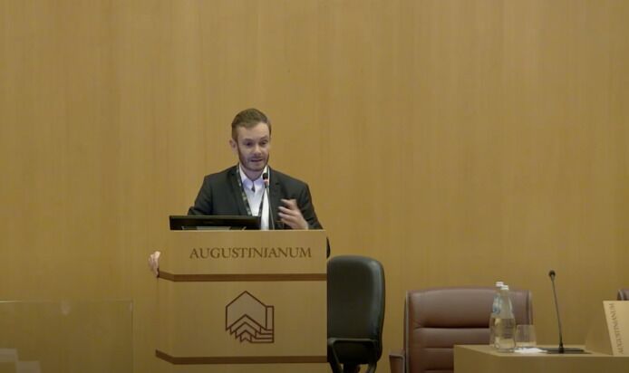 A man in a dark suit stands at a podium labeled 'AUGUSTINIANUM' giving a presentation. He gesticulates with his left hand while speaking. The background is a plain, light-colored wall, and there are some empty chairs and a water bottle to his right.