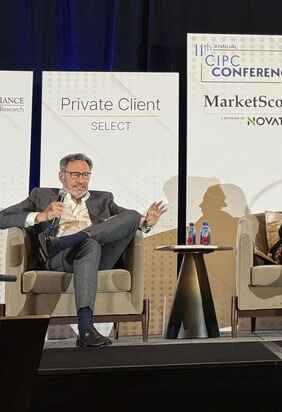 A man in a gray suit is seated on a beige chair, participating in a panel discussion at the 11th annual CIPC Conference. Behind him are banners displaying 'Private Client Select' and 'MarketScope.' A small table with water bottles is beside him.